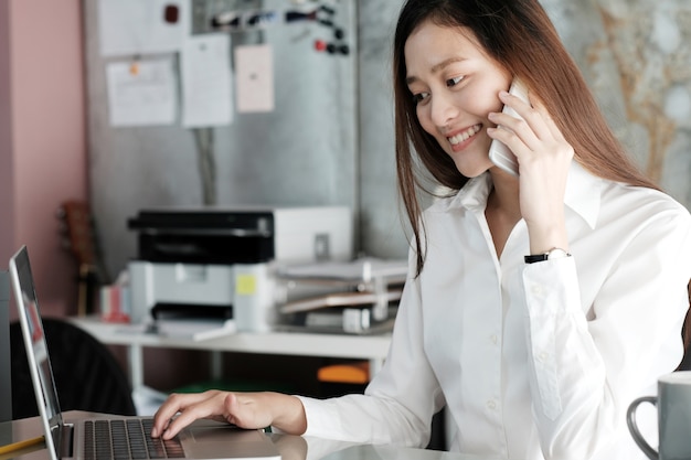 Junge Geschäftsfrau, die intelligentes Telefon spricht und mit Laptop arbeitet