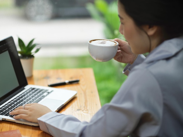 Junge Geschäftsfrau, die Kaffee trinkt und nippt, während sie an ihrem Laptop arbeitet und ihren Präsentationsprozess entwirft.