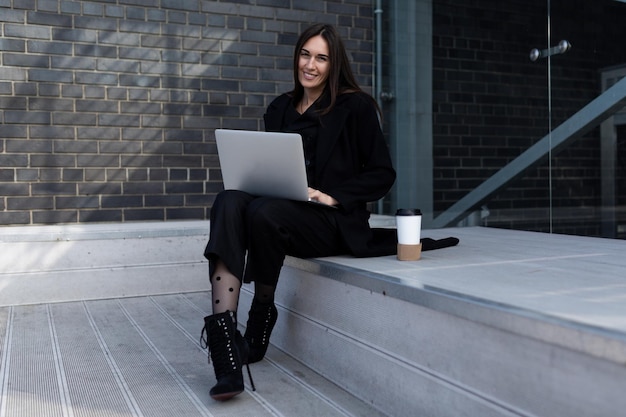 Junge Geschäftsfrau, die online an einem Laptop arbeitet, mit einer Tasse Kaffee, die auf den Stufen in der Nähe sitzt