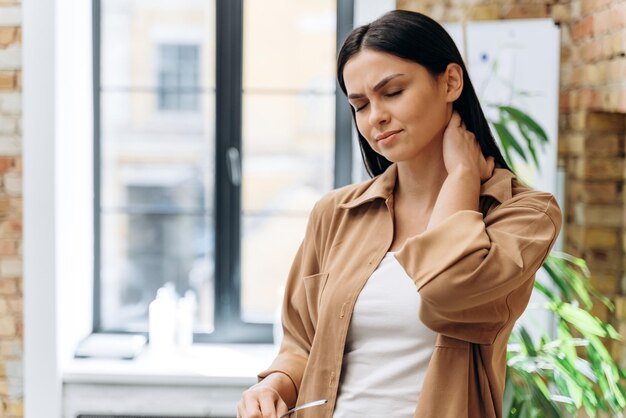 Junge Geschäftsfrau, die steifen Nacken berührt und massiert, um Muskelschmerzen nach sitzender Computerarbeit in falscher Haltung zu lindern. Frau mit Fibromyalgie bei der Arbeit