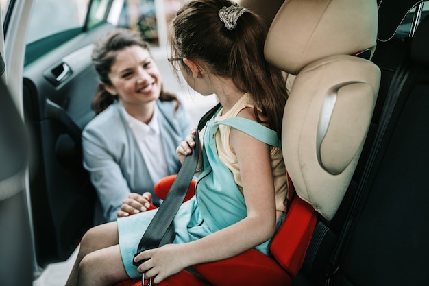 Junge Geschäftsfrau hilft ihrer Tochter beim Anschnallen im Auto, während das Mädchen auf einem Sicherheitskindersitz sitzt.