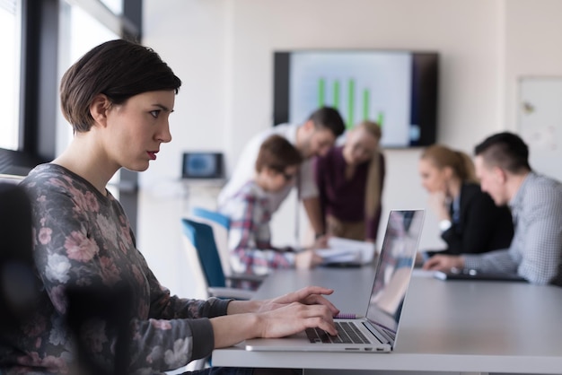 junge geschäftsfrau im modernen startbüroinnenraum, die an laptop-computer arbeitet, verschwommenes team bei der besprechung, personengruppe im hintergrund