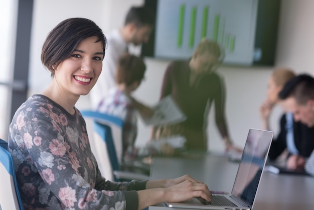 junge geschäftsfrau im modernen startbüroinnenraum, die an laptop-computer arbeitet, verschwommenes team bei der besprechung, personengruppe im hintergrund