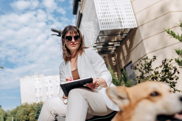 Junge Geschäftsfrau im weißen Anzug, die auf einem Elektroroller sitzt und an einem digitalen Tablet arbeitet, während sie mit dem walisischen Corgi-Pembroke-Hund in der Stadt spazieren geht