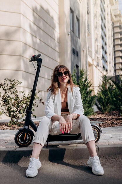 Junge Geschäftsfrau im weißen Anzug sitzt auf einem Elektroroller, während sie in der Stadt spazieren geht und in die Kamera schaut