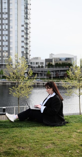 Junge Geschäftsfrau in Anzug, freiberufliche Arbeitnehmerin, die mit dem Laptop im Freien im Park arbeitet, Büroangestellte, die auf einem Rasen in einem Stadtstraßenpark auf einem modernen städtischen Hintergrund sitzt, Arbeitsplatz im Freien