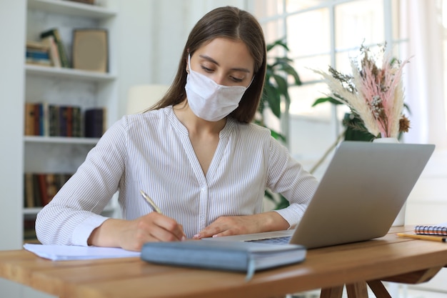 Junge Geschäftsfrau in einer medizinischen Schutzmaske arbeitet während der Selbstisolation und Quarantäne von zu Hause am Laptop.