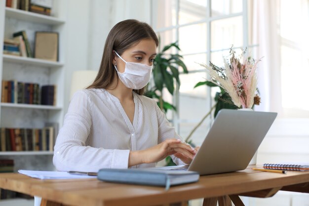 Junge Geschäftsfrau in einer medizinischen Schutzmaske arbeitet während der Selbstisolation und Quarantäne von zu Hause am Laptop.