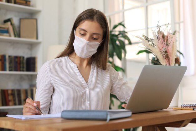 Junge Geschäftsfrau in einer medizinischen Schutzmaske arbeitet während der Selbstisolation und Quarantäne von zu Hause am Laptop.