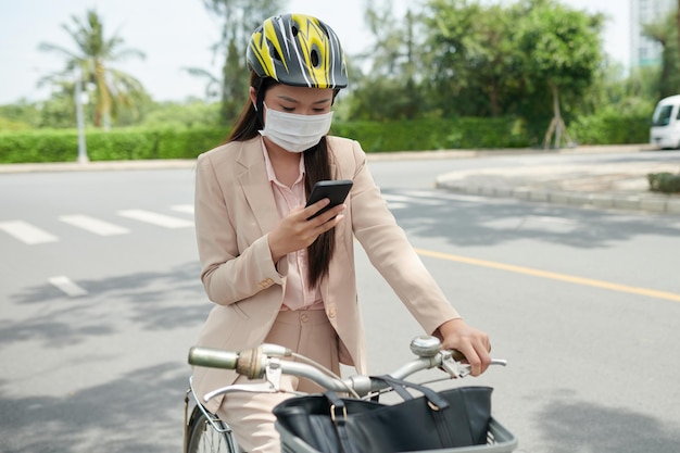 Junge Geschäftsfrau in Schutzmaske sitzt auf dem Fahrrad und überprüft Benachrichtigungen auf dem Smartphone