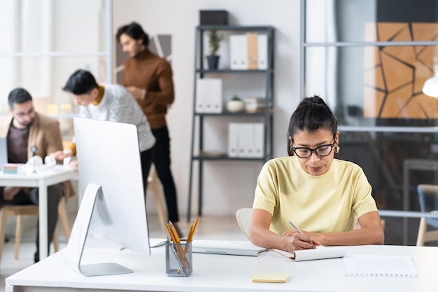 Junge Geschäftsfrau mit Brille sitzt an ihrem Arbeitsplatz mit Computer und macht sich während ihrer Arbeit im Büro Notizen im Notizbuch