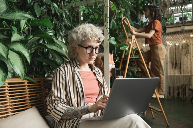 Junge Geschäftsfrau mit Brille sitzt auf dem Sofa und arbeitet online am Laptop im Blumenladen mit ihrer Kollegin im Hintergrund