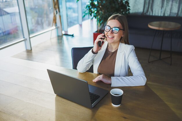 Junge Geschäftsfrau mit Brille und Freizeitkleidung sitzt an einem Holztisch mit Laptop und einer Tasse Kaffee und macht Hausaufgaben auf einem Laptop