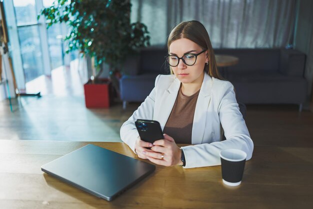 Junge Geschäftsfrau mit Brille und Freizeitkleidung sitzt an einem Holztisch mit Laptop und einer Tasse Kaffee und macht Hausaufgaben auf einem Laptop