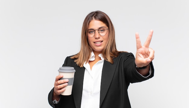 Junge Geschäftsfrau mit einem Kaffee, der lächelt und glücklich, sorglos und positiv aussieht, Sieg oder Frieden mit einer Hand gestikulierend