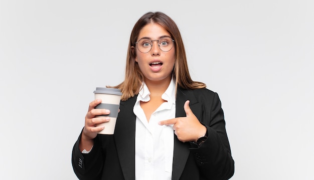 Junge Geschäftsfrau mit einem Kaffee, der sich glücklich, überrascht und stolz fühlt und mit einem aufgeregten, erstaunten Blick auf sich selbst zeigt