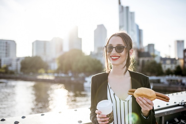 Junge Geschäftsfrau mit einem Snack mit traditioneller Frankfurter Wurst und Kaffee auf der Brücke mit Wolkenkratzern im Hintergrund in Frankfurt während des Sonnenuntergangs