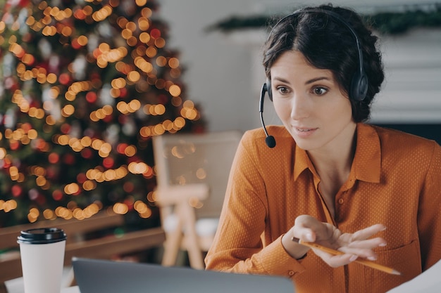 Junge Geschäftsfrau mit Headset im Gespräch mit Kollegen während eines Online-Meetings im Büro zu Weihnachten
