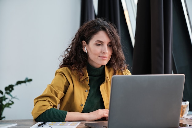 Junge Geschäftsfrau mit Kopfhörern, die etwas auf dem Laptop beobachtet, während sie an ihrem Arbeitsplatz sitzt