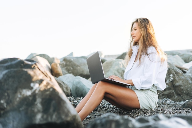 Junge Geschäftsfrau mit langen Haaren in einem weißen Hemd, die bei Sonnenaufgang an der Küste am Laptop arbeitet