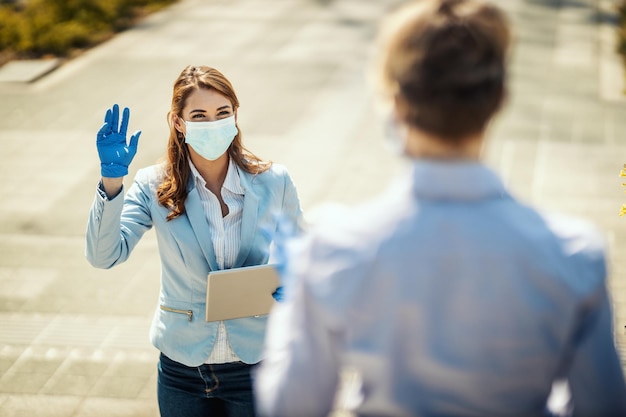 Junge geschäftsfrau mit schutzmaske im gesicht wird arbeiten, um ihrer freundin während der covid19-pandemie hallo zu sagen.