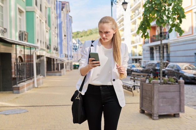 Junge Geschäftsfrau mit Smartphone, die entlang der Stadtstraße geht, weibliche Lesung, Textnachricht eingeben, E-Mail suchen