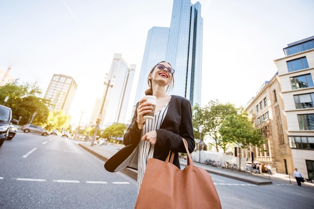 Junge Geschäftsfrau mit Tasche und Kaffee beim Überqueren der Straße im modernen Stadtteil Frankfurt