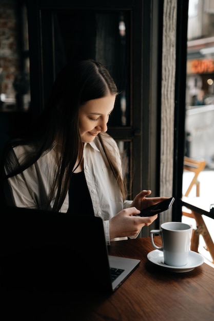 Junge Geschäftsfrau schreibt Nachricht auf ihrem Telefon, trinkt schwarzen Kaffee und lächelt