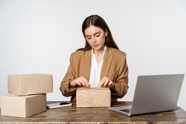 Junge Geschäftsfrau, selbstständige Frau, die im Büro in der Nähe von Laptop sitzt und Pakete verpackt, die Kunden verpacken...