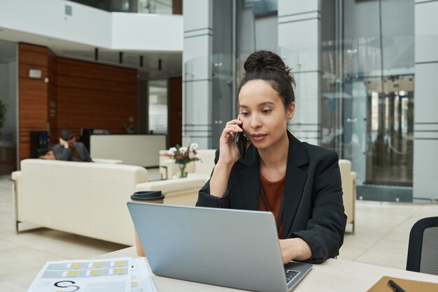 Junge Geschäftsfrau sitzt am Tisch, arbeitet am Laptop und telefoniert im Büro mit dem Handy