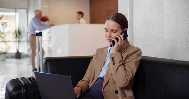 Junge Geschäftsfrau sitzt in der Lobby des Hotels, telefoniert und benutzt einen Laptop