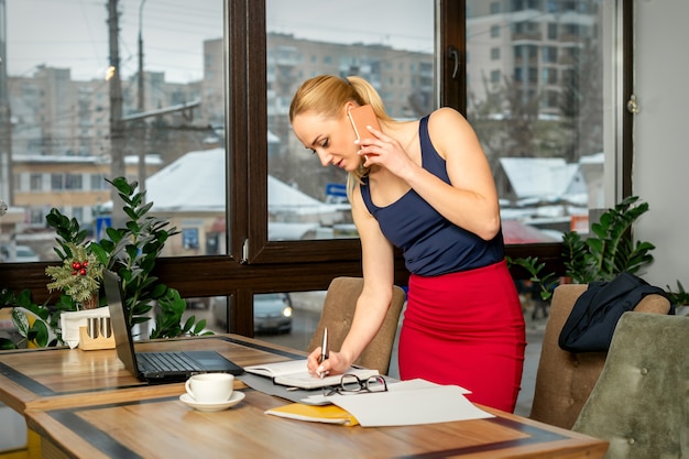 Junge Geschäftsfrau spricht am Telefon und schreibt Notizen, die im Büro oder im Café stehen