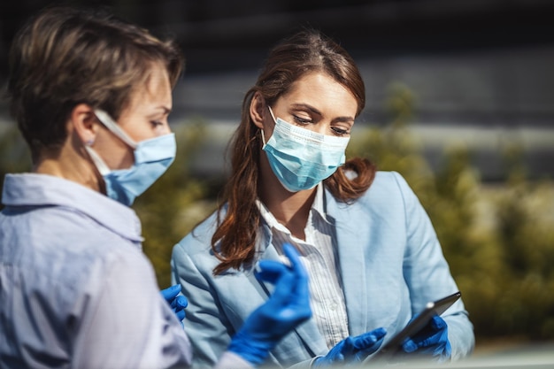 Junge Geschäftskollegen mit Schutzmasken im Gesicht analysieren das Projekt im Freien und sprechen darüber, während sie ein digitales Tablet verwenden.