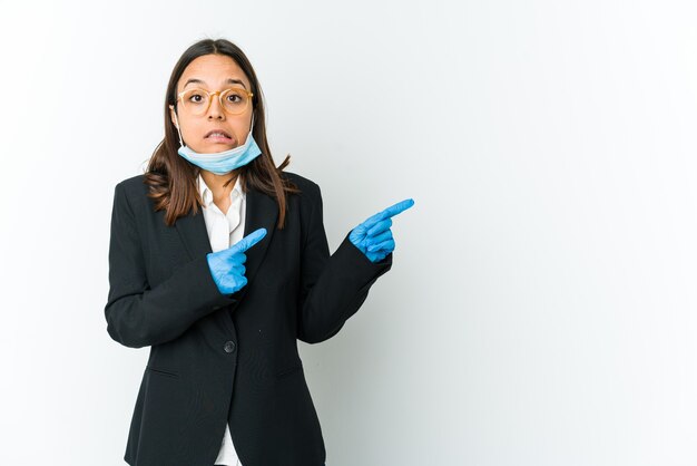 Junge Geschäftslateinfrau, die eine Maske trägt, um vor Covid zu schützen, isoliert auf weißem schockiertem Zeigen mit Zeigefingern zu einem Kopienraum.