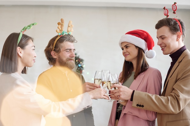 Junge Geschäftsleute in Weihnachtsstirnbändern, die mit Champagnerflöten klirren, während sie auf die erfolgreiche Arbeit im nächsten Jahr anstoßen