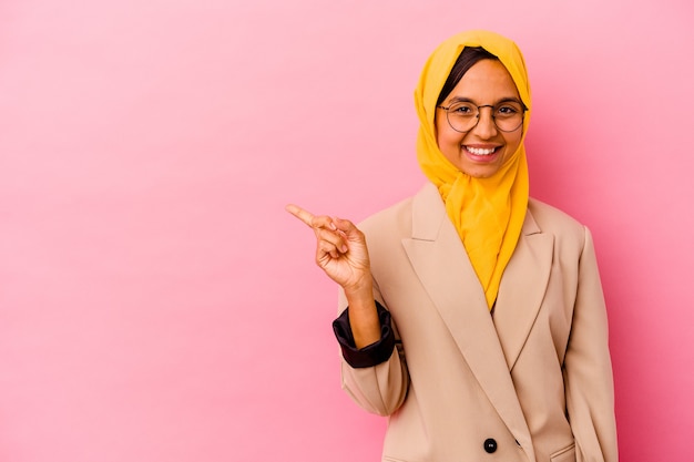 Junge Geschäftsmuslimfrau lokalisiert auf rosa Wand, die lächelt und beiseite zeigt und etwas an der leeren Stelle zeigt.