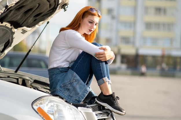 Junge gestresste Fahrerin in der Nähe eines kaputten Autos mit geknallter Motorhaube, die auf Hilfe wartet