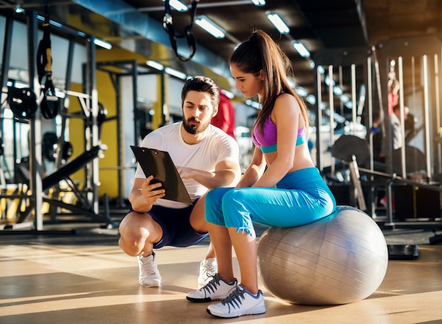 Junge gesunde aktive Frau, die auf dem Gymnastikball sitzt und mit einem Personal Trainer über einen Übungsplan berät.