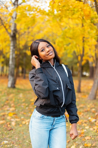 Junge glamouröse, stilvolle schwarze kanadische Frau in modischer Freizeitjacke mit Blue Jeans, die im Herbstpark mit leuchtend gelbem Laub spaziert