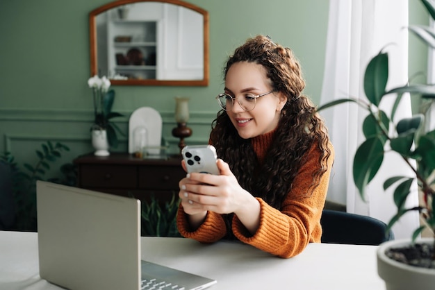 Junge, glücklich lächelnde, hübsche Frau, die am Tisch sitzt und ihr Smartphone in der Hand hält und ein modernes Mobiltelefon nutzt