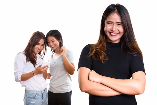 Junge glückliche asiatische Frau, die mit gekreuzten Armen mit zwei glücklichen Freunden auf der Rückseite unter Verwendung des Mobiltelefons zusammen lächelt