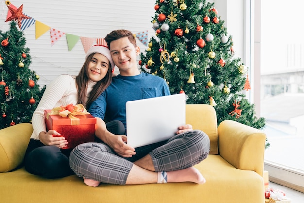 Junge glückliche asiatische Paare, die zusammen Videoanruf auf einem Laptop mit Freunden oder Eltern sitzen, danke für ein Geschenk, das während der Weihnachtsferien erhalten wurde. Paar schaut in die Kamera