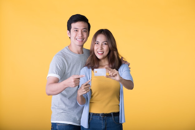 Junge glückliche asiatische Paarfinger, die auf Kreditkarte lokalisiert auf gelber Wand zeigen