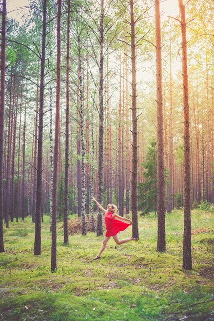 Junge glückliche blonde Frau im roten Kleid, die im Kiefernwald tanzt