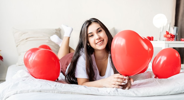 Junge glückliche brünette Frau, die im Bett mit roten herzförmigen Luftballons liegt