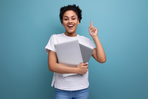 Junge glückliche brünette Frau mit Afro-Haaren in einem weißen Tanktop hält einen Laptop für die Arbeit in ihren Händen