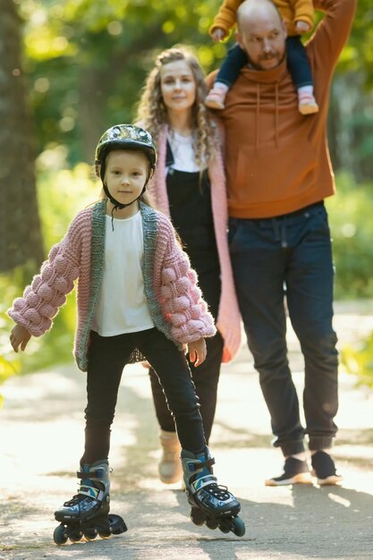 Junge, glückliche Elternfamilie und zwei kleine Kinder, die im Wald spazieren gehen, ein Mädchen, das auf den Rollen Schlittschuh läuft