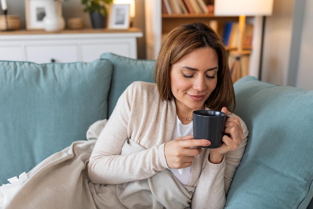 Foto junge, glückliche, entspannte frau, die ruhige momente zu hause mit einer tasse tee oder kaffee genießt, lächelnde frau, die einen becher mit heißem getränk hält, auf dem sofa im wohnzimmer ruht und davon träumt, sich von allem zu entspannen