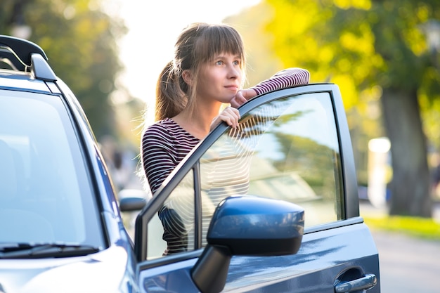 Junge glückliche Fahrerin, die im Sommer in der Nähe ihres Autos auf einer Stadtstraße steht. Reiseziele und Transportkonzept.