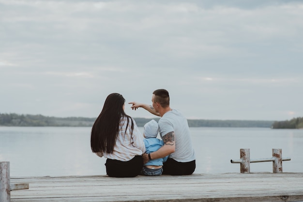 Junge glückliche Familie, die auf dem Pier im Sommer am Wasser sitzt